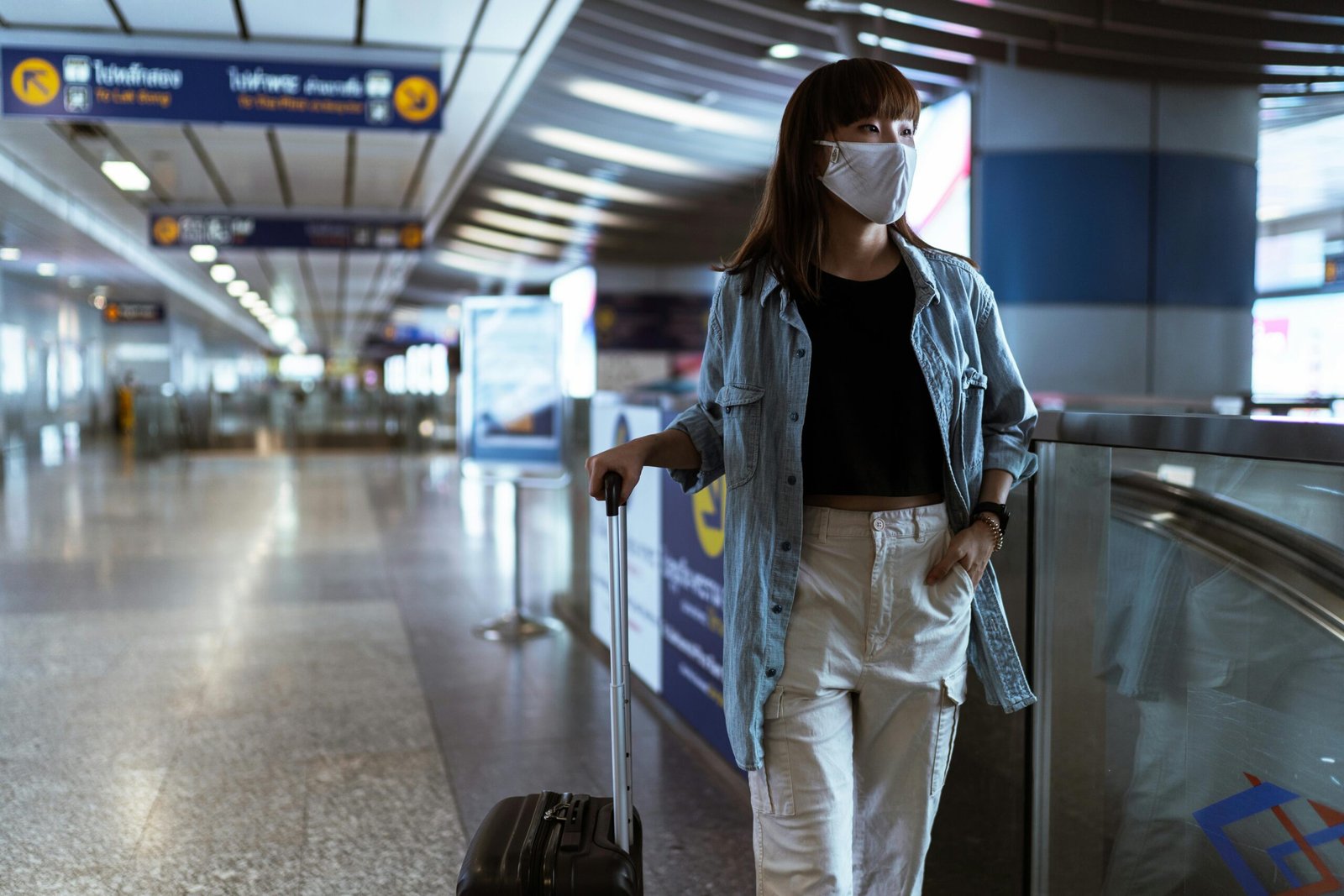 Woman wearing mask and denim jacket in airport terminal with luggage.
